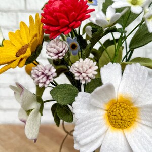 Puede incluir: Un vibrante ramo de flores de fieltro en un jarr&oacute;n de cristal transparente. El arreglo incluye un gran crisantemo rojo, un girasol amarillo, margaritas blancas y peque&ntilde;as flores rosas y azules. Hojas y tallos verdes completan la colorida exhibici&oacute;n.
