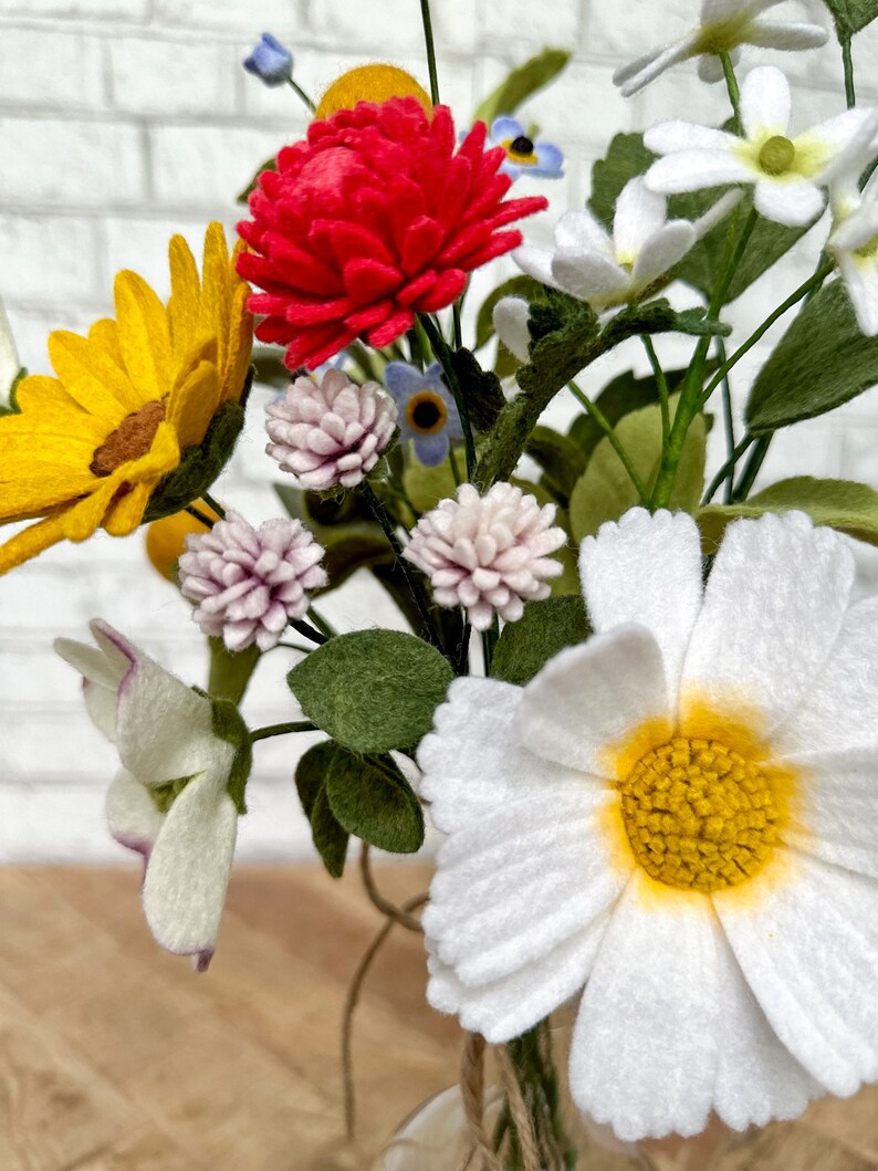 Puede incluir: Un vibrante ramo de flores de fieltro en un jarr&oacute;n de cristal transparente. El arreglo incluye un gran crisantemo rojo, un girasol amarillo, margaritas blancas y peque&ntilde;as flores rosas y azules. Hojas y tallos verdes completan la colorida exhibici&oacute;n.