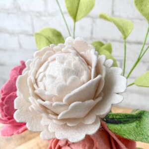 May include: Close-up of felt flowers in shades of pink and cream, with green felt leaves. The flowers have detailed petal construction. The background is a white brick wall. The arrangement is a craft item.