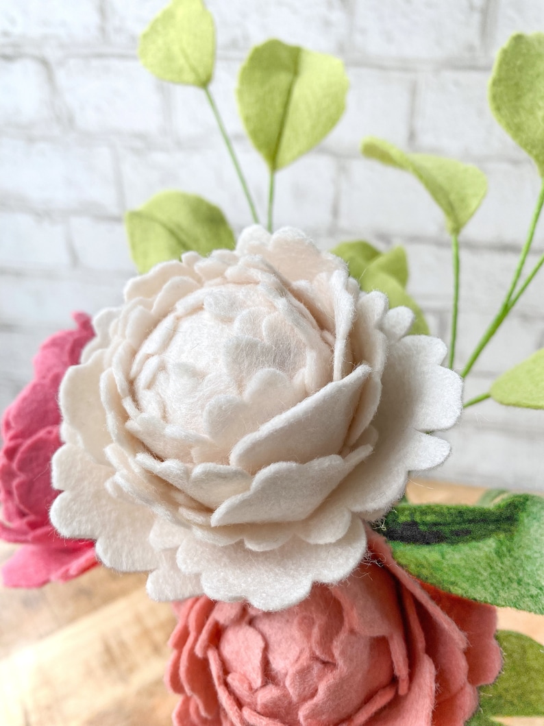 May include: Close-up of felt flowers in shades of pink and cream, with green felt leaves. The flowers have detailed petal construction. The background is a white brick wall. The arrangement is a craft item.