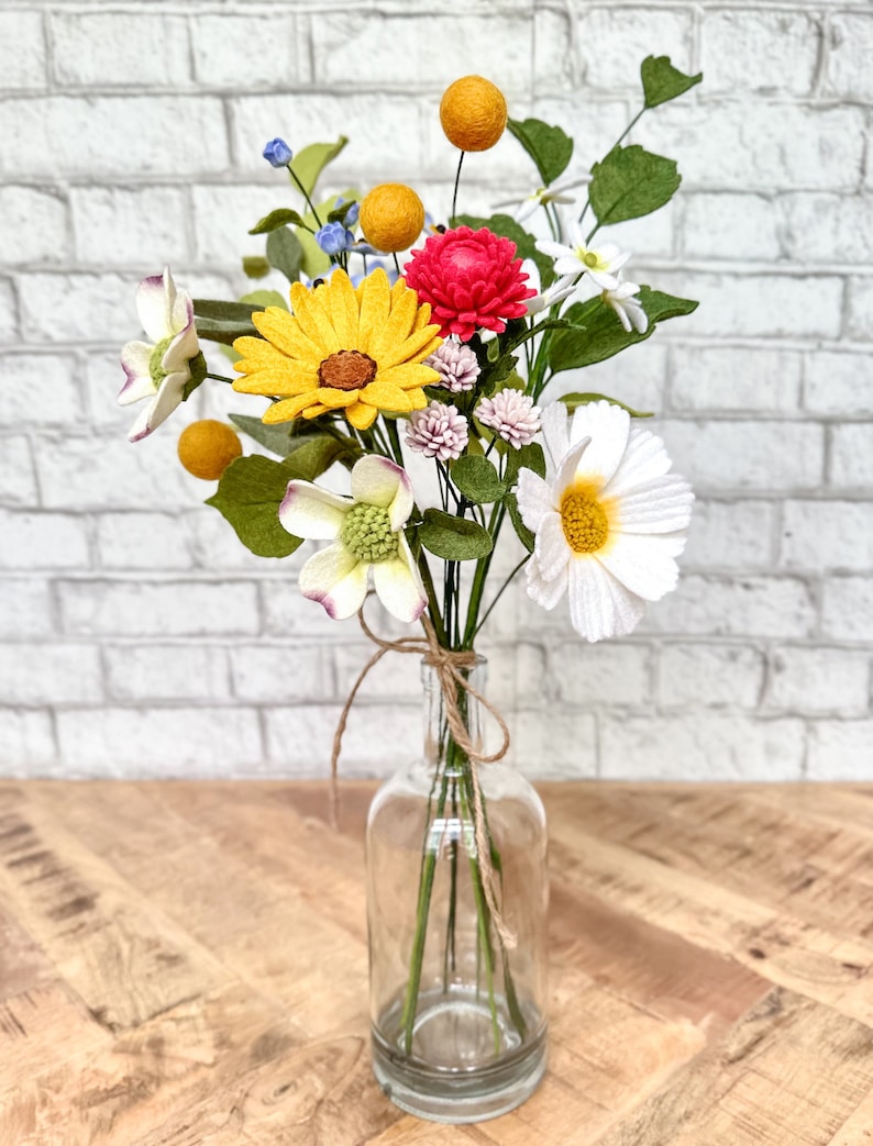 Puede incluir: Un ramo de flores de fieltro de colores en una botella de vidrio transparente atada con hilo. El arreglo incluye girasoles amarillos, crisantemos rojos, margaritas blancas y hojas verdes.