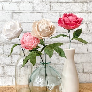 Op de afbeelding: Vier vilten pioenrozen in wit, roze en rood, met groene bladeren, tentoongesteld in glazen en keramische vazen. De vazen staan op een houten oppervlak tegen een witte bakstenen muur.
