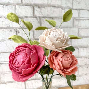 May include: A bouquet of felt peonies in a clear glass vase. The arrangement features three large, textured flowers in shades of pink and peach, with a cream-colored bloom. Green leaves and stems complete the floral display.