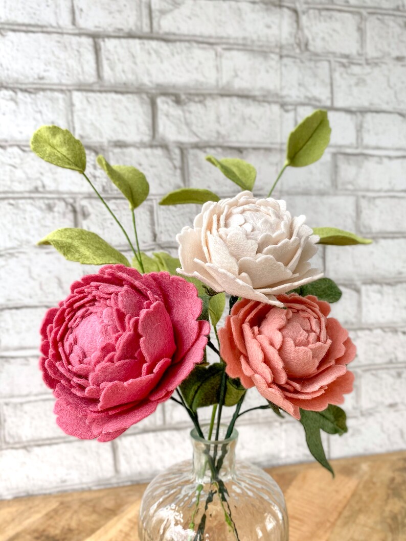 May include: A bouquet of felt peonies in a clear glass vase. The arrangement features three large, textured flowers in shades of pink and peach, with a cream-colored bloom. Green leaves and stems complete the floral display.