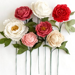 Op de afbeelding: Een verzameling handgemaakte vilten pioenrozen in verschillende kleuren, waaronder wit, rood, roze en cr&egrave;me. Elke bloem heeft groene bladeren en een lange groene steel. De opstelling staat tegen een witte achtergrond.