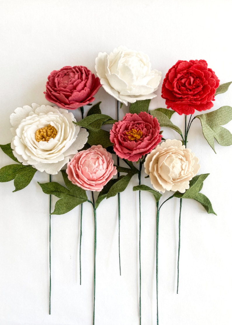 Op de afbeelding: Een verzameling handgemaakte vilten pioenrozen in verschillende kleuren, waaronder wit, rood, roze en cr&egrave;me. Elke bloem heeft groene bladeren en een lange groene steel. De opstelling staat tegen een witte achtergrond.