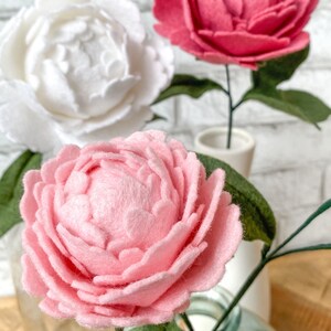 Op de afbeelding: Close-up van drie vilten pioenrozen in wit, roze en rood. De bloemen hebben groene stelen en bladeren en worden tentoongesteld in glazen en keramische vazen tegen een witte bakstenen muur.