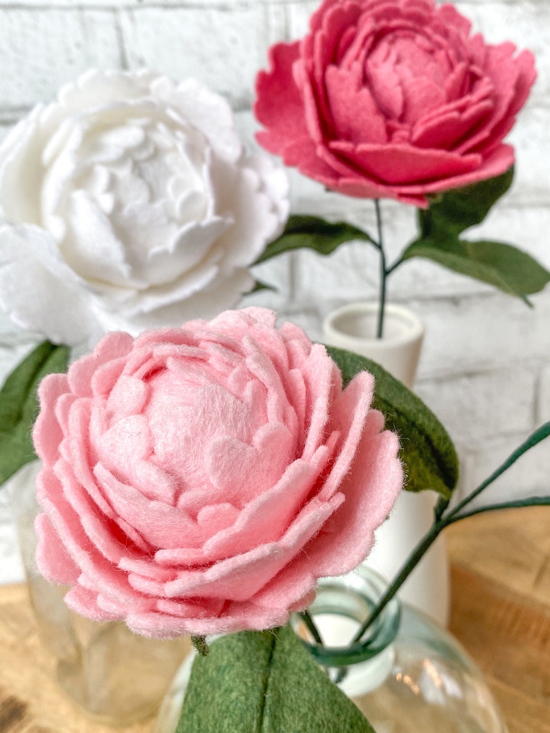 Op de afbeelding: Close-up van drie vilten pioenrozen in wit, roze en rood. De bloemen hebben groene stelen en bladeren en worden tentoongesteld in glazen en keramische vazen tegen een witte bakstenen muur.