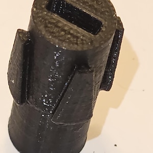 May include: A black, cylindrical object with a rectangular aperture at the top and two protruding rectangular shapes on the sides. The object has a textured surface, appearing to be made of a solid material.