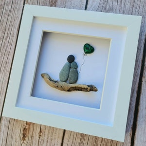 May include: A framed pebble art piece depicting two figures holding each other, sitting on a piece of driftwood. A green heart-shaped balloon is floating above them.