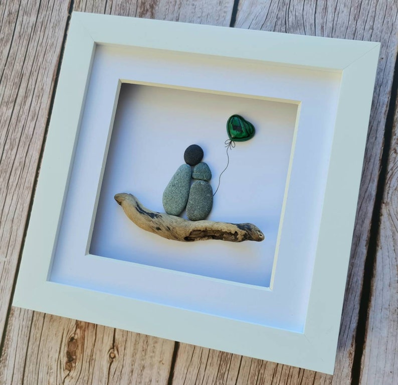 May include: A framed pebble art piece depicting two figures holding each other, sitting on a piece of driftwood. A green heart-shaped balloon is floating above them.