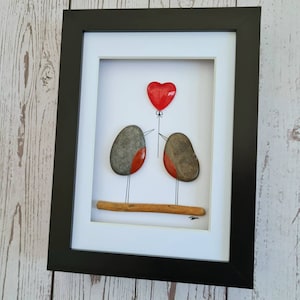 Op de afbeelding: Ingelijst kiezelkunstwerk met twee vogels die een rode hartballon vasthouden. De vogels zijn gemaakt van grijze en rode stenen, en de ballon is van rood glas. Het kunstwerk is geplaatst tegen een witte achtergrond.
