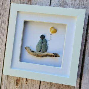 May include: A framed pebble art piece depicting two figures holding hands, sitting on a piece of driftwood. A yellow heart-shaped balloon is floating above them.