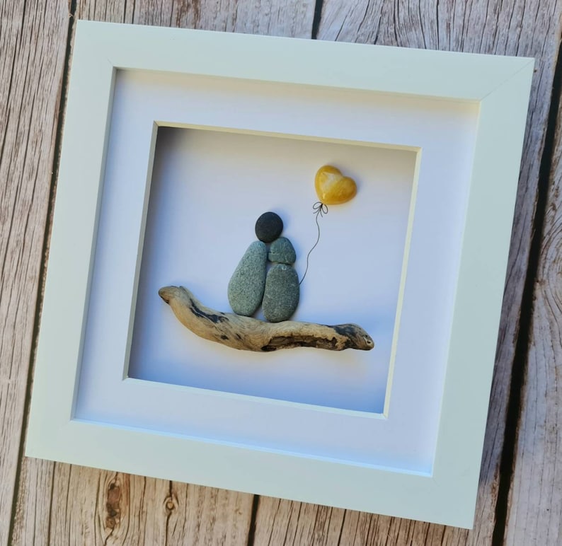 May include: A framed pebble art piece depicting two figures holding hands, sitting on a piece of driftwood. A yellow heart-shaped balloon is floating above them.