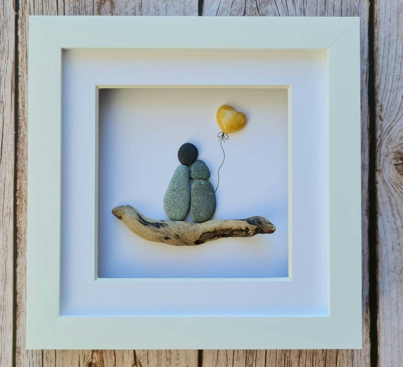 May include: A framed pebble art piece depicting two figures holding hands, sitting on a piece of driftwood with a yellow heart balloon floating above them.