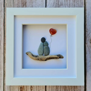May include: A framed pebble art piece depicting two gray stones representing a couple sitting on a piece of driftwood. A red heart shaped stone is floating above them on a string.