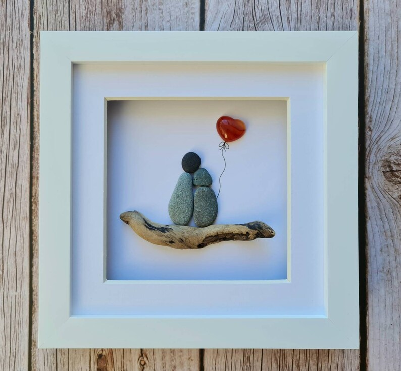 May include: A framed pebble art piece depicting two gray stones representing a couple sitting on a piece of driftwood. A red heart shaped stone is floating above them on a string.