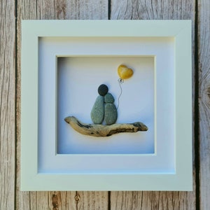 May include: A framed pebble art piece depicting two figures holding hands, sitting on a driftwood branch. A yellow heart-shaped balloon floats above them.
