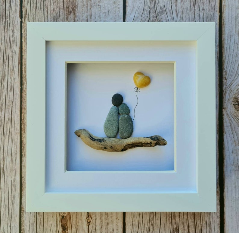 May include: A framed pebble art piece depicting two figures holding hands, sitting on a driftwood branch. A yellow heart-shaped balloon floats above them.