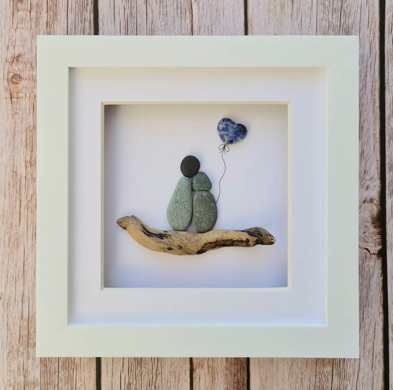May include: A framed pebble art piece depicting two figures holding hands, sitting on a piece of driftwood. A blue heart-shaped balloon floats above them.