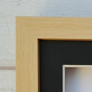 May include: Close-up of a natural wood picture frame with a black mat and a white inner bevel. The frame has a simple, clean design.