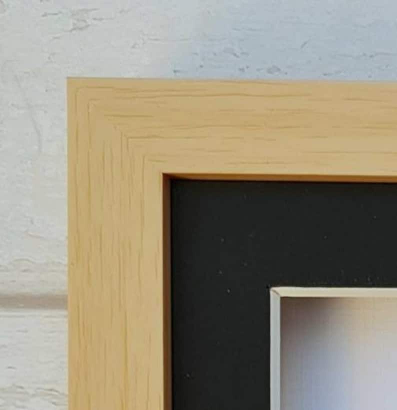 May include: Close-up of a natural wood picture frame with a black mat and a white inner bevel. The frame has a simple, clean design.