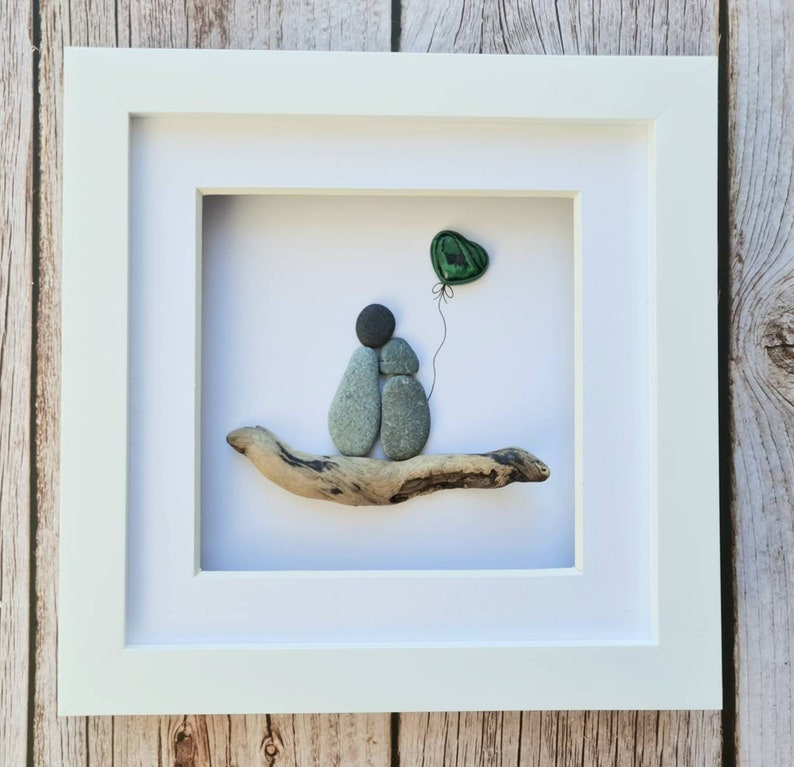 May include: A framed pebble art piece depicting two figures holding hands, sitting on a piece of driftwood with a green heart-shaped balloon floating above them.