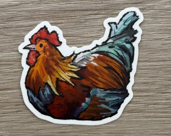 May include: A colorful sticker featuring a detailed illustration of a rooster. The rooster has a red comb and wattle, with brown, orange, and blue feathers. The sticker is outlined in white and is on a wooden surface.