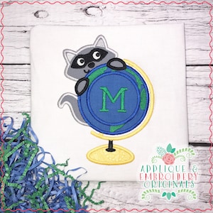 May include: Embroidered design of a raccoon holding a globe with the letter M on it. The globe is blue and green with a yellow stand. The design is on a white background.