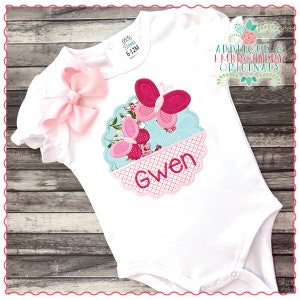 May include: White baby bodysuit with a pink bow, a blue and pink floral circle with two pink butterflies and the name "Gwen" embroidered in pink.