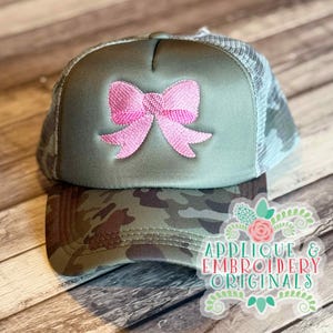 May include: A camouflage trucker hat with a pink bow embroidered on the front. The hat has a mesh back and a curved brim. The text "APPLIQUE & EMBROIDERY ORIGINALS" is visible on the lower right.