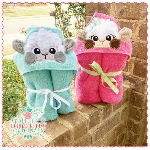 May include: Two hooded towels with embroidered sheep faces. The towel on the left is turquoise with a gray sheep face. The towel on the right is pink with a brown sheep face.