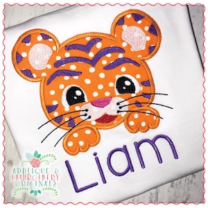 May include: Orange and purple embroidered tiger applique with white polka dots. The tiger is looking at the viewer with a friendly expression. The name "Liam" is embroidered in purple below the tiger.