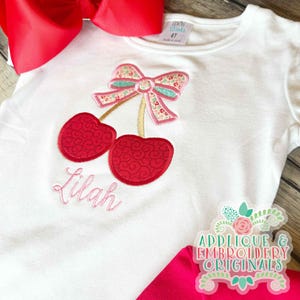 Peut inclure: T-shirt blanc à manches courtes avec une application de cerises. Les cerises sont rouges avec un motif décoratif, et les tiges et le nœud sont floraux. Le nom "Lilah" est brodé en rose en dessous. Un grand nœud rouge est en arrière-plan.