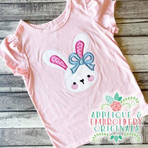 Puede incluir: Camiseta rosa claro de manga corta con un aplique de conejo blanco, orejas de lunares rosas y un lazo azul. El conejo tiene mejillas rosas y ojos negros. El texto "APPLIQUE & EMBROIDERY ORIGINALS" está en la parte inferior.