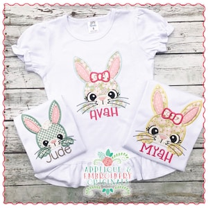 Pode incluir: Três camisas brancas com rostos de coelho bordados e nomes. Os rostos de coelho são verdes, rosa e amarelos com laços rosa. Os nomes são Jude, Avah e Myah. As camisas estão à venda pela Applique & Embroidery Originals.