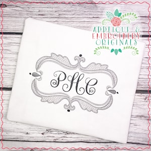 May include: Embroidered monogram "PHC" in a black script font within a silver ornate frame. The frame has a decorative, scalloped edge. The design is on a white fabric background.