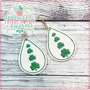 May include: A pair of white teardrop-shaped earrings with green shamrock embroidery. The earrings have gold hooks.