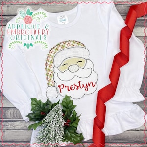 May include: White long-sleeved shirt with a red and green plaid Santa applique. The applique has a white beard and the name "Prestyln" is embroidered in red below the beard.