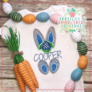 May include: White onesie with a blue and gray bunny applique, the name "COOPER" embroidered below, and a green and blue checkered pattern on the bunny's head. The onesie is surrounded by colorful Easter eggs and carrots.