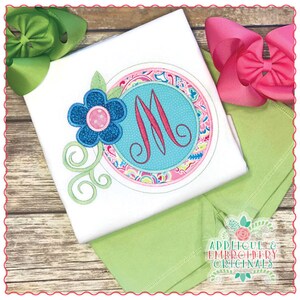 May include: A white t-shirt with a green and pink floral applique featuring a blue flower and a turquoise circle with a pink letter M in the center. The shirt is on a wooden surface with a green and pink bow on each side.