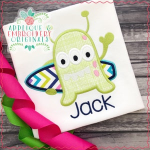 May include: Green three-eyed monster with a toothy grin, riding a surfboard with colorful stripes. The monster is wearing a green and white checkered pattern. The name "Jack" is embroidered in blue below the monster.