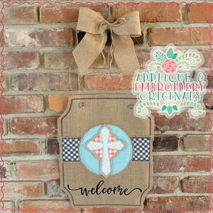 May include: A burlap welcome sign with a white cross in a blue circle, a black and white checkered ribbon, and the word "welcome" in cursive. The sign is hanging on a brick wall with a burlap bow.
