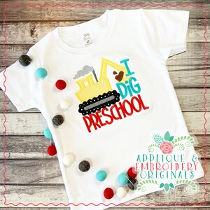 May include: White t-shirt with an embroidered design of a yellow excavator with the text "I Dig Preschool".