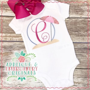 May include: White onesie with a pink and blue beach umbrella and the letter C embroidered on it. The onesie has a circle around the design. The text "Applique & Embroidery Originals" is on the bottom of the onesie.