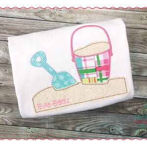 May include: A white t-shirt with an embroidered design of a pink bucket with sand, a blue shovel, and the name "Evie Begu" in brown thread.