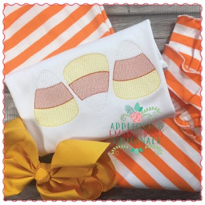 May include: Three embroidered candy corn designs on a white t-shirt. The candy corn is yellow, orange, and white. The t-shirt is on top of an orange and white striped fabric.