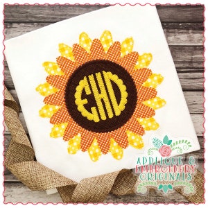 Puede incluir: Una camiseta blanca con una aplicación de girasol amarillo y naranja. El girasol tiene un centro marrón con un monograma amarillo "EHD" en su interior. La camiseta está sobre una superficie de madera con una cinta de arpillera marrón.