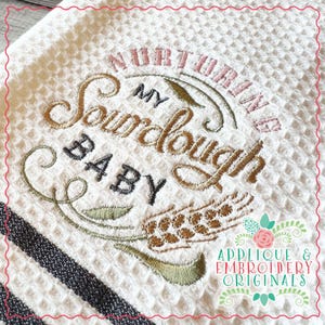 May include: White kitchen towel with a black and gray stripe design. The towel has embroidered text that reads "Nourishing My Sourdough Baby" with a wheat stalk design.