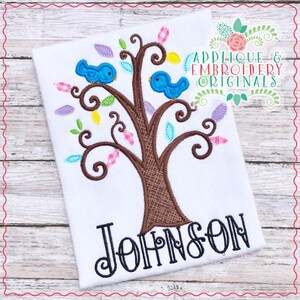 Può includere: Una maglietta bianca con un albero marrone con uccelli blu e foglie colorate ricamate. Il testo "Johnson" è ricamato sotto l'albero.
